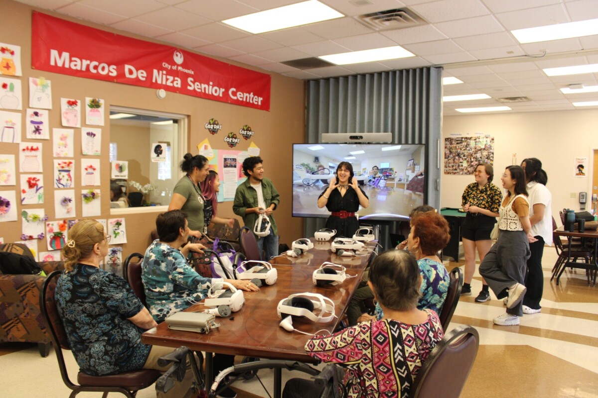 A group of seniors sitting around a table with VR headsets in front of them, looking at a very funny presenter telling a joke.